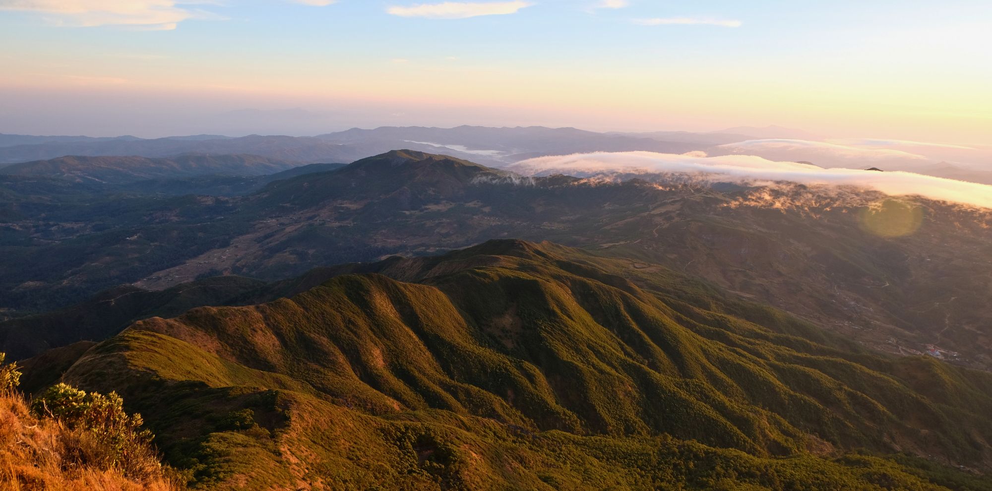 A panoramic view of rolling mountains covered in lush greenery, bathed in warm sunlight during sunset. Wispy clouds hover over the peaks, with layers of hills fading into the background.