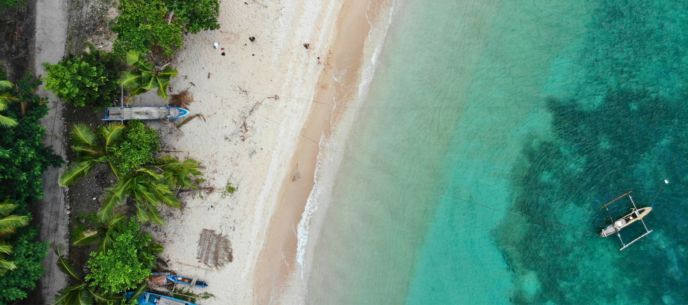 Aerial view of a sandy beach lined with palm trees and small fishing boats. The turquoise water gently laps at the shore, creating a serene coastal scene.
