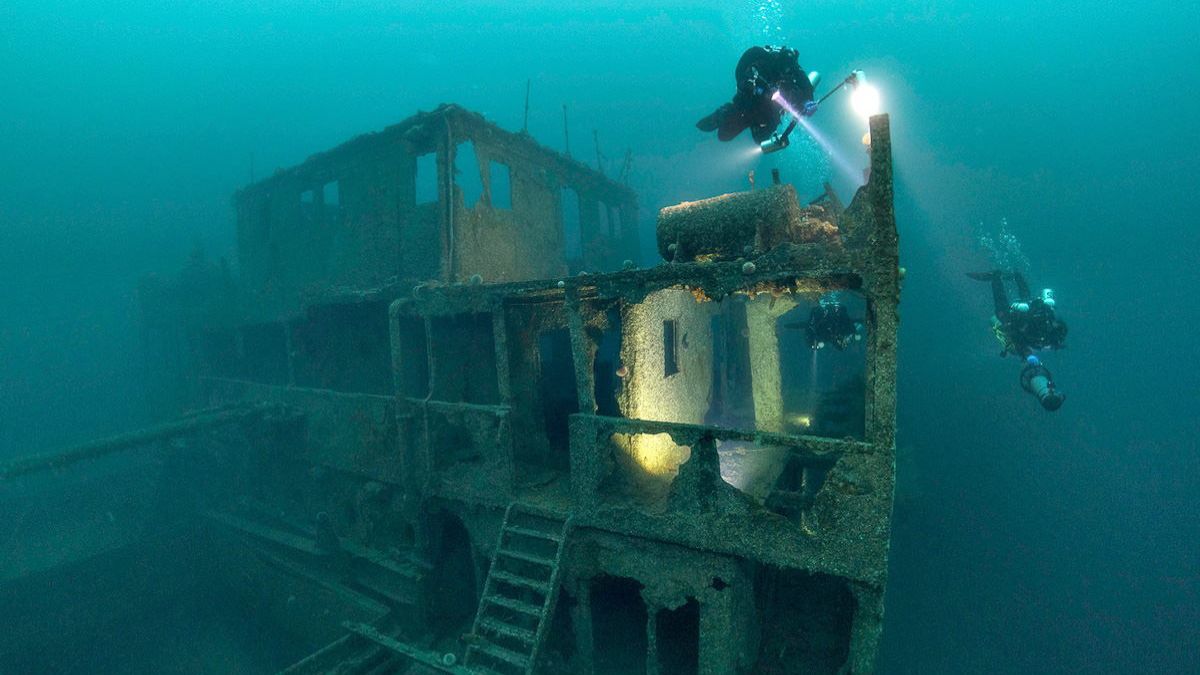 Divers explore a sunken shipwreck, illuminating the dark underwater environment with their torches. The structure shows signs of rust and decay, with large openings and remnants of rooms visible. Bubbles rise as they navigate through the eerie, blue depths.