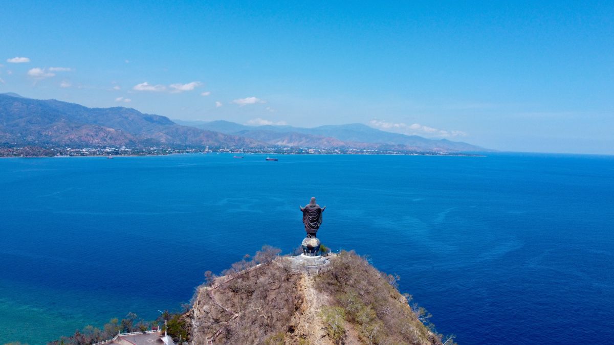 A large statue stands on a cliff overlooking the ocean, surrounded by mountains in the background. The sky is clear with a few clouds, and the water is calm, showcasing shades of blue.