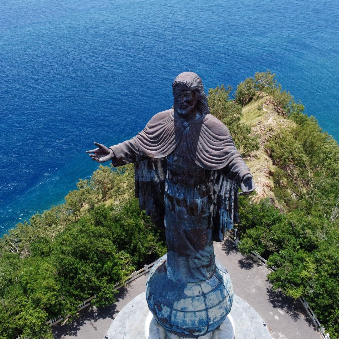 A large statue of Christ with outstretched arms stands on a rocky cliff, overlooking a vibrant blue ocean. Lush greenery surrounds the base of the statue, creating a serene atmosphere.