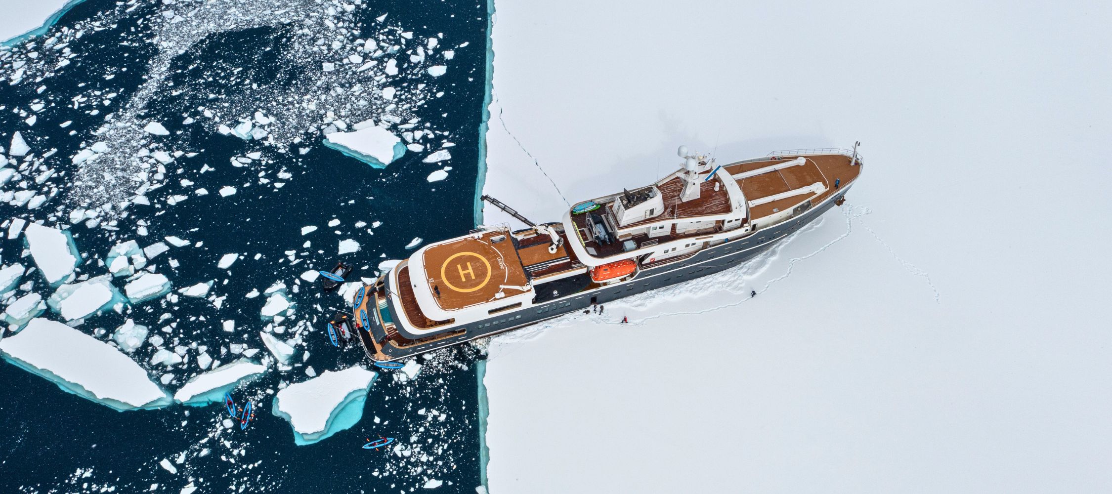 A large research vessel navigates through icy waters, with icebergs surrounding it on a stark white landscape. The ship features a helipad on its top deck and appears to be breaking through the ice.