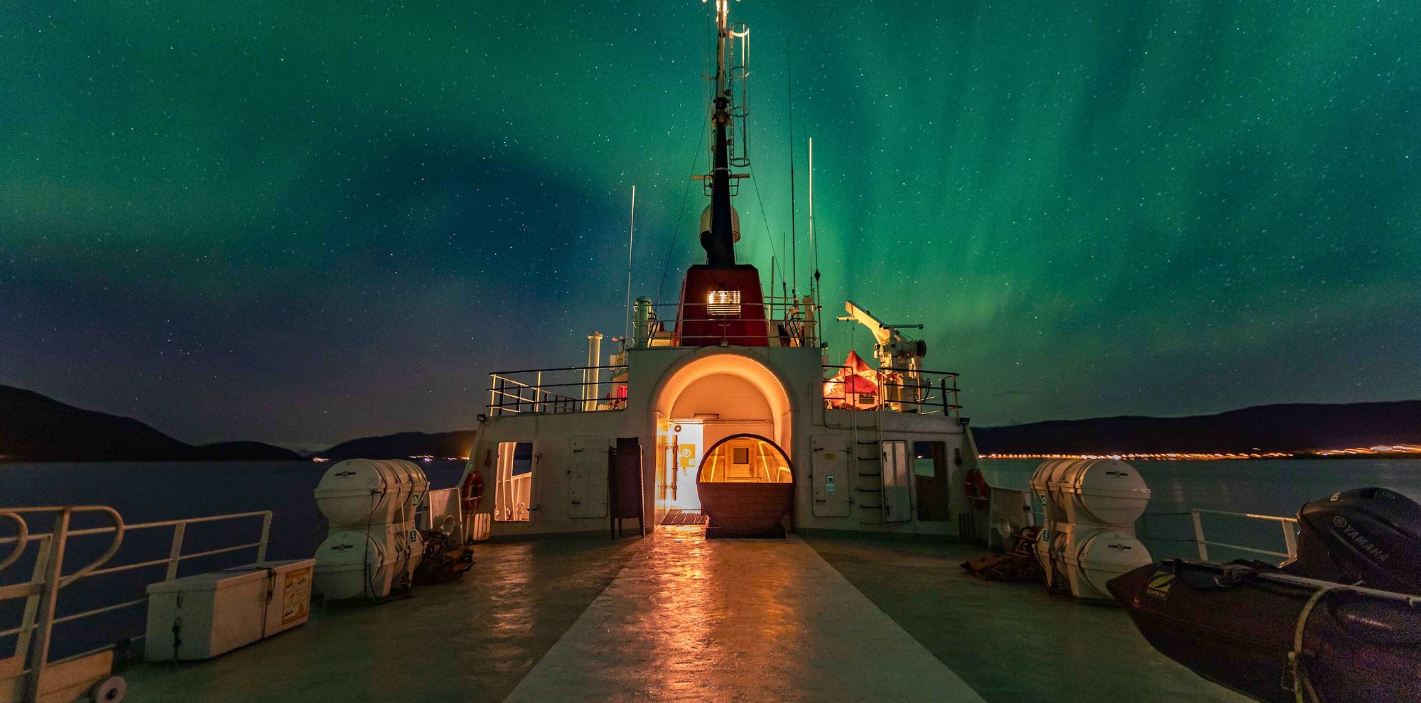 A ship's deck illuminated by the glow of the northern lights, with bright green and subtle purple hues in the sky. The ship's structure is visible in the foreground, leading towards an illuminated doorway. The surrounding water reflects the vibrant colors of the aurora borealis.