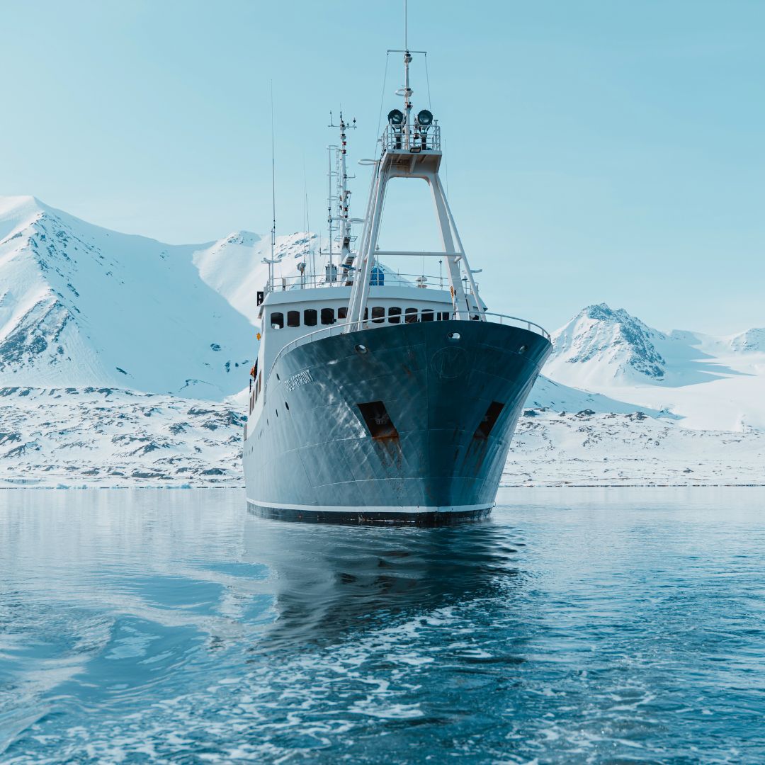 A large ship with a blue hull is anchored in icy waters, surrounded by snow-covered mountains under a clear blue sky.