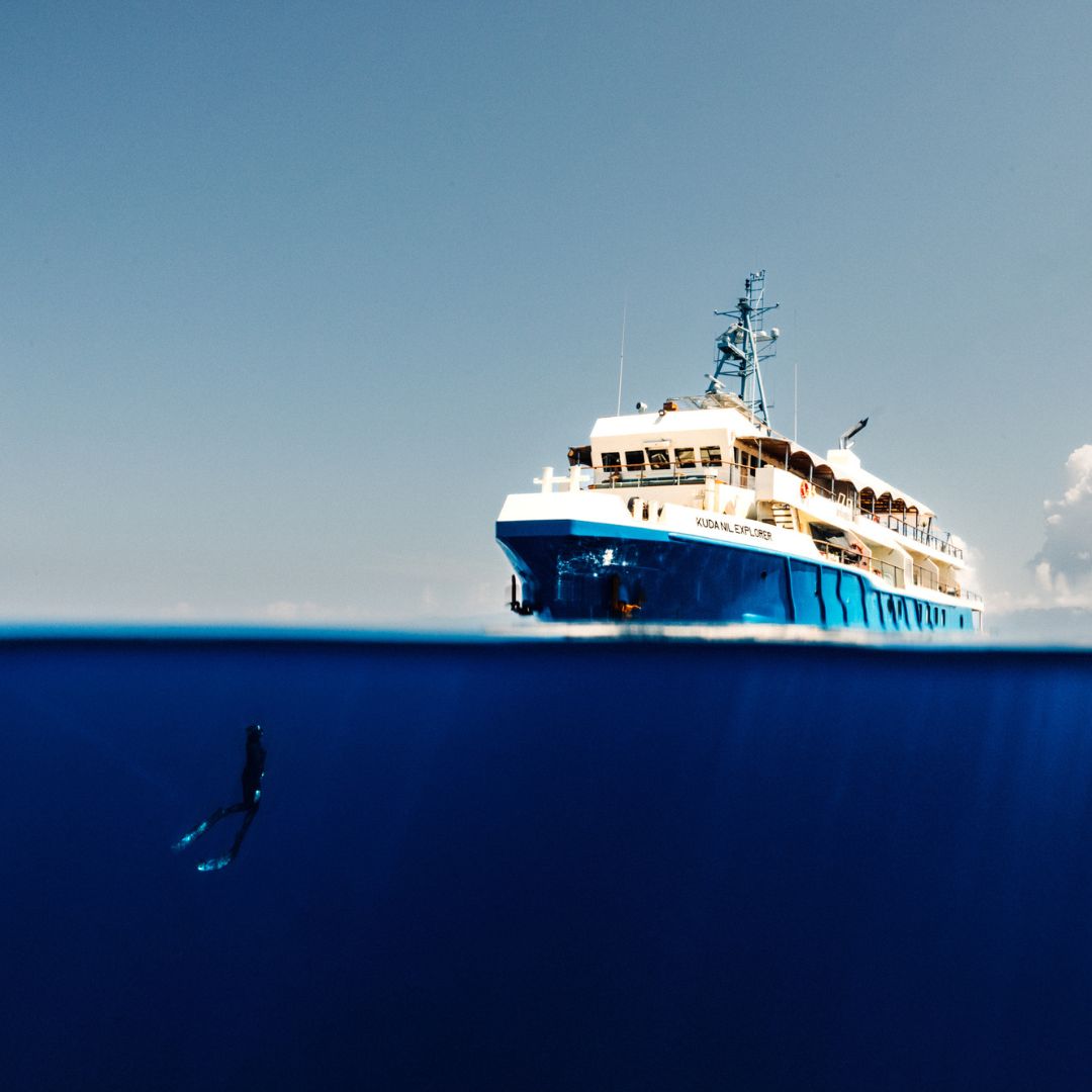A diver swims beneath the surface of the ocean, with a large boat visible above water against a clear blue sky.