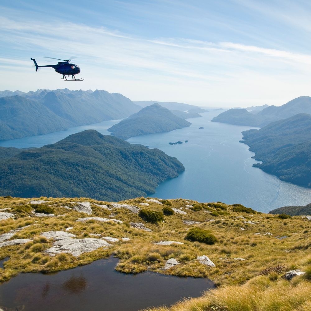 A blue helicopter flies over a vast landscape of lush green mountains and deep blue fjords.