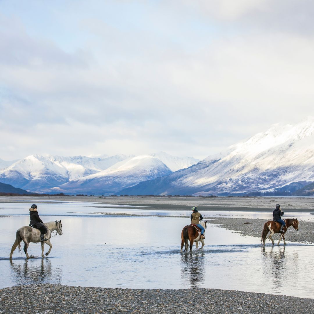 Three people riding horses through a shallow river with snow-covered mountains in the background.
