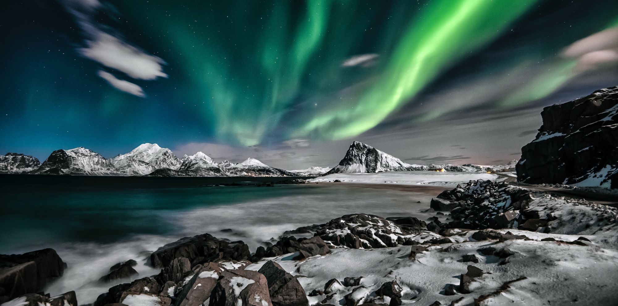 Vibrant green Aurora Borealis sweeps across a starry night sky over a snowy coastline with jagged mountains and dark water.