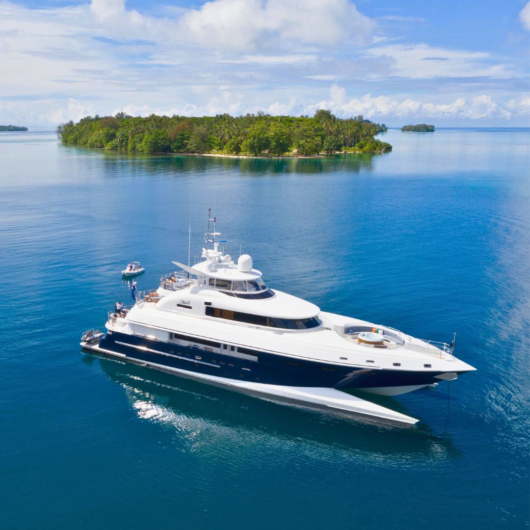 A luxurious yacht cruising in calm blue waters, with a tropical island lush with greenery in the background under a partly cloudy sky.