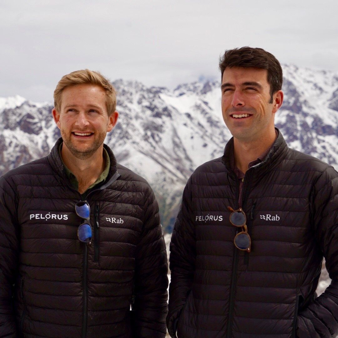 Two men wearing black jackets with logos stand against a backdrop of snow-capped mountains. They are smiling, with a cloudy sky above.