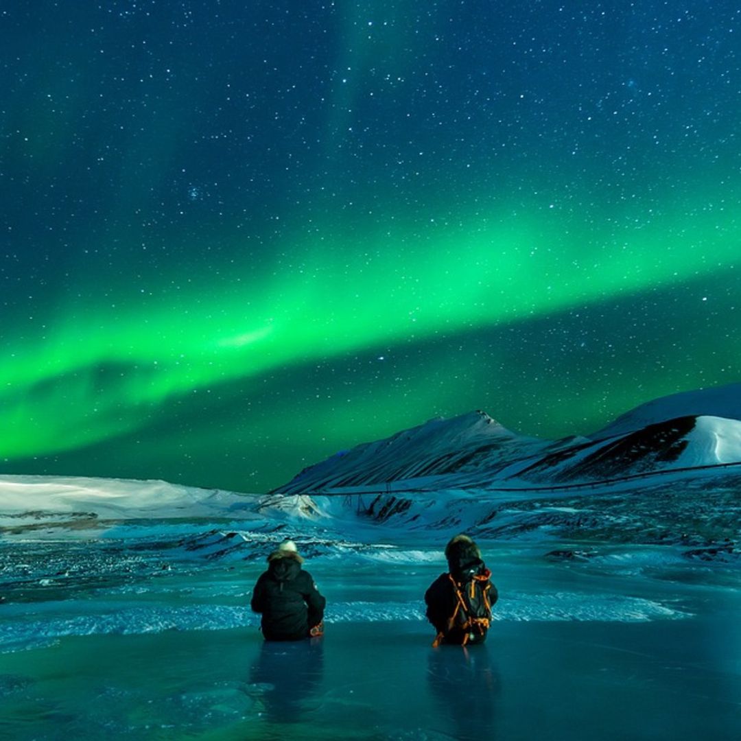 Two individuals sit on a frozen landscape, gazing up at the vibrant green lights of the Northern Lights against a starry sky, with snow-capped mountains in the background.