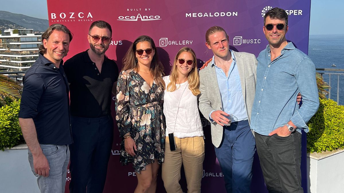 A group of six people poses together outdoors, smiling in front of a colorful backdrop featuring various brand logos. The scene has a scenic ocean view in the background, and the individuals are dressed in smart casual attire.