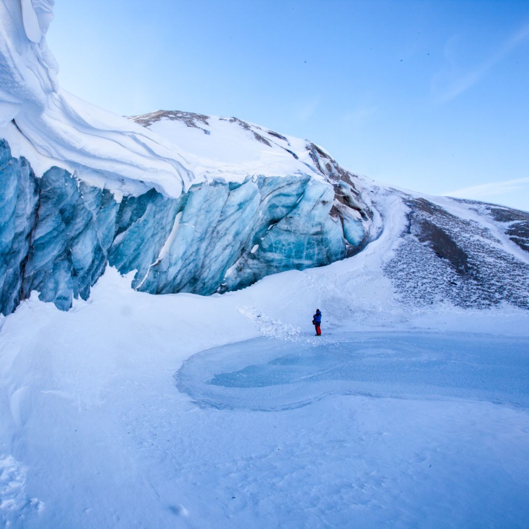 A person stands on a snowy landscape near a large, blue-tinted glacier, surrounded by snow-covered terrain and a clear blue sky.