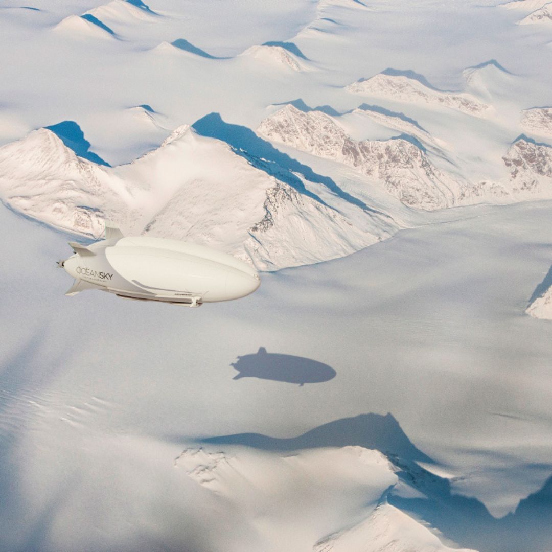A sleek, white airship glides over a snow-covered landscape with mountains in the background, casting a shadow on the icy terrain below.