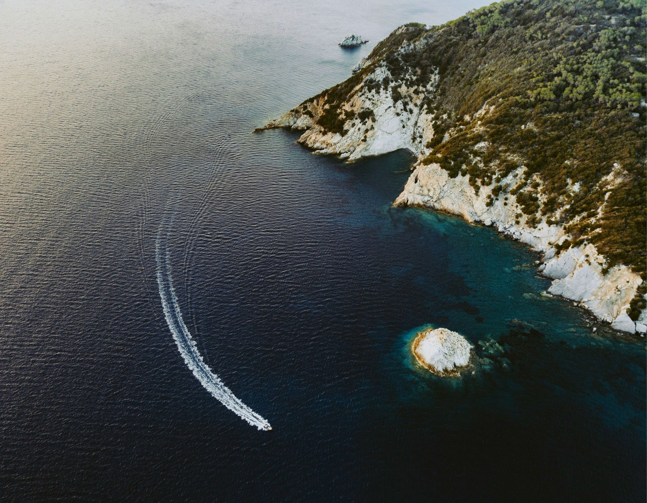 Aerial view of a boat leaving a white wake in dark blue water near a rugged, tree-covered coastline.