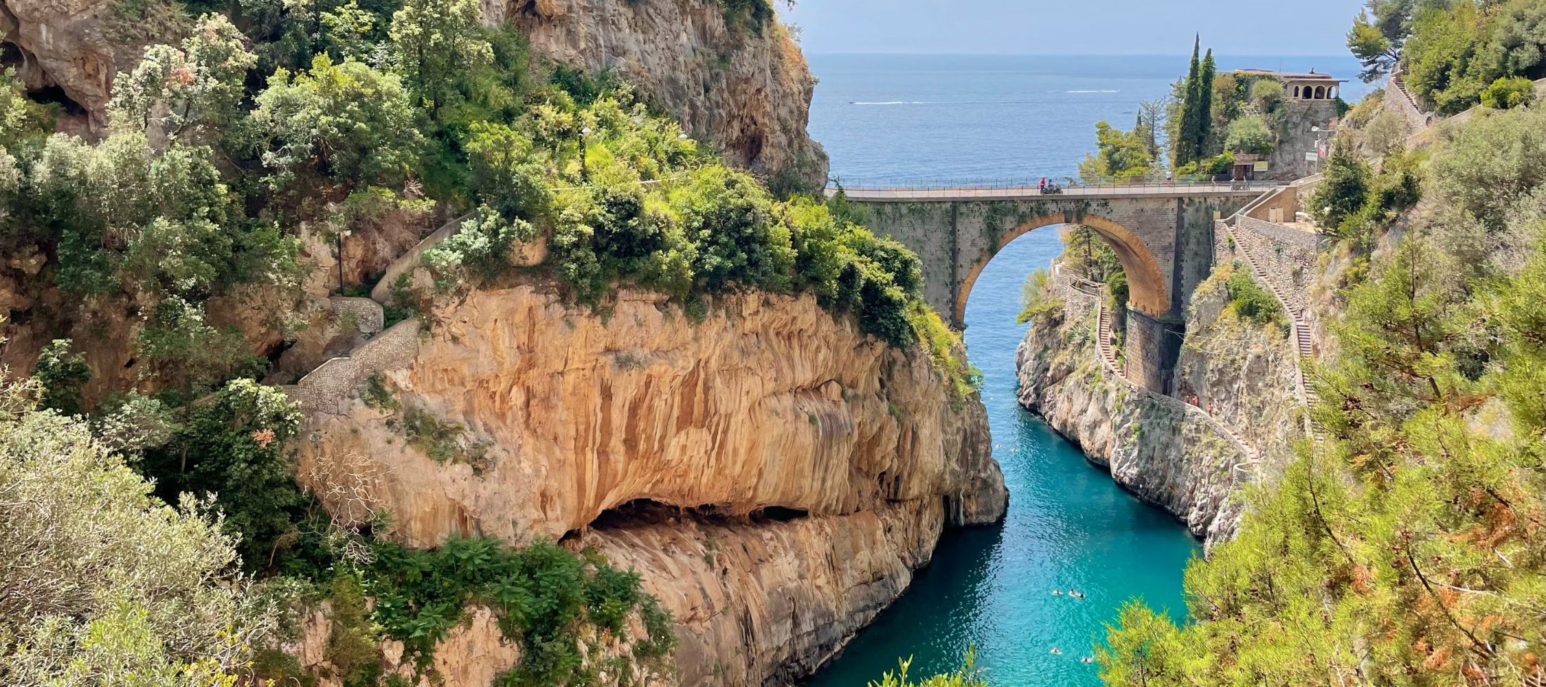 A stone arch bridge spans a narrow turquoise sea inlet between steep, craggy cliffs covered in green vegetation.
