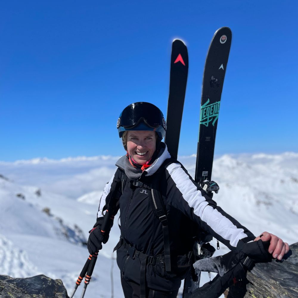 A skier stands on a mountain peak, smiling confidently while holding ski poles and skis against a backdrop of snowy slopes and a clear blue sky.