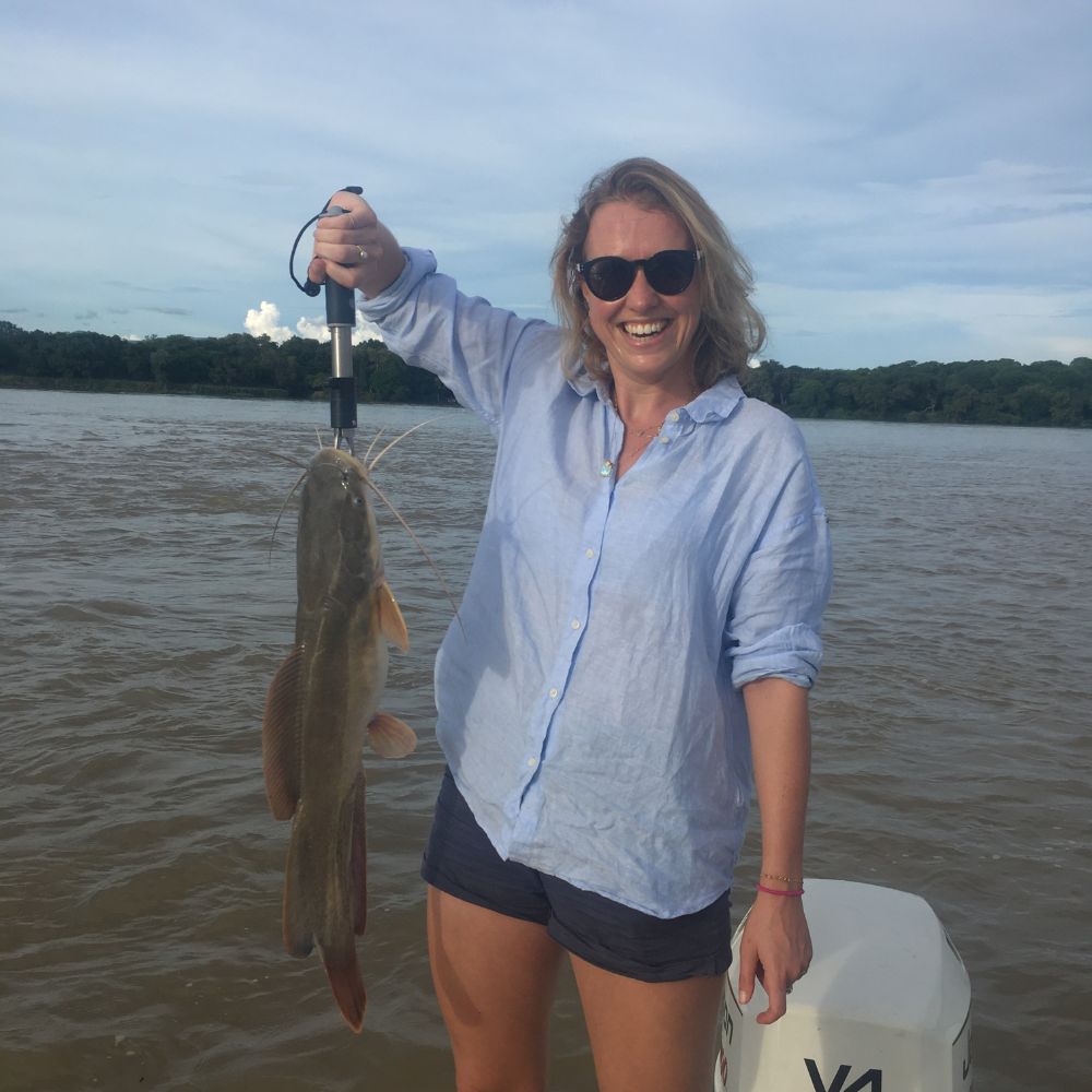 A smiling woman wearing sunglasses and a light blue shirt holds a large catfish by its tail over a boat on a river. Green trees and a cloudy sky are visible in the background.