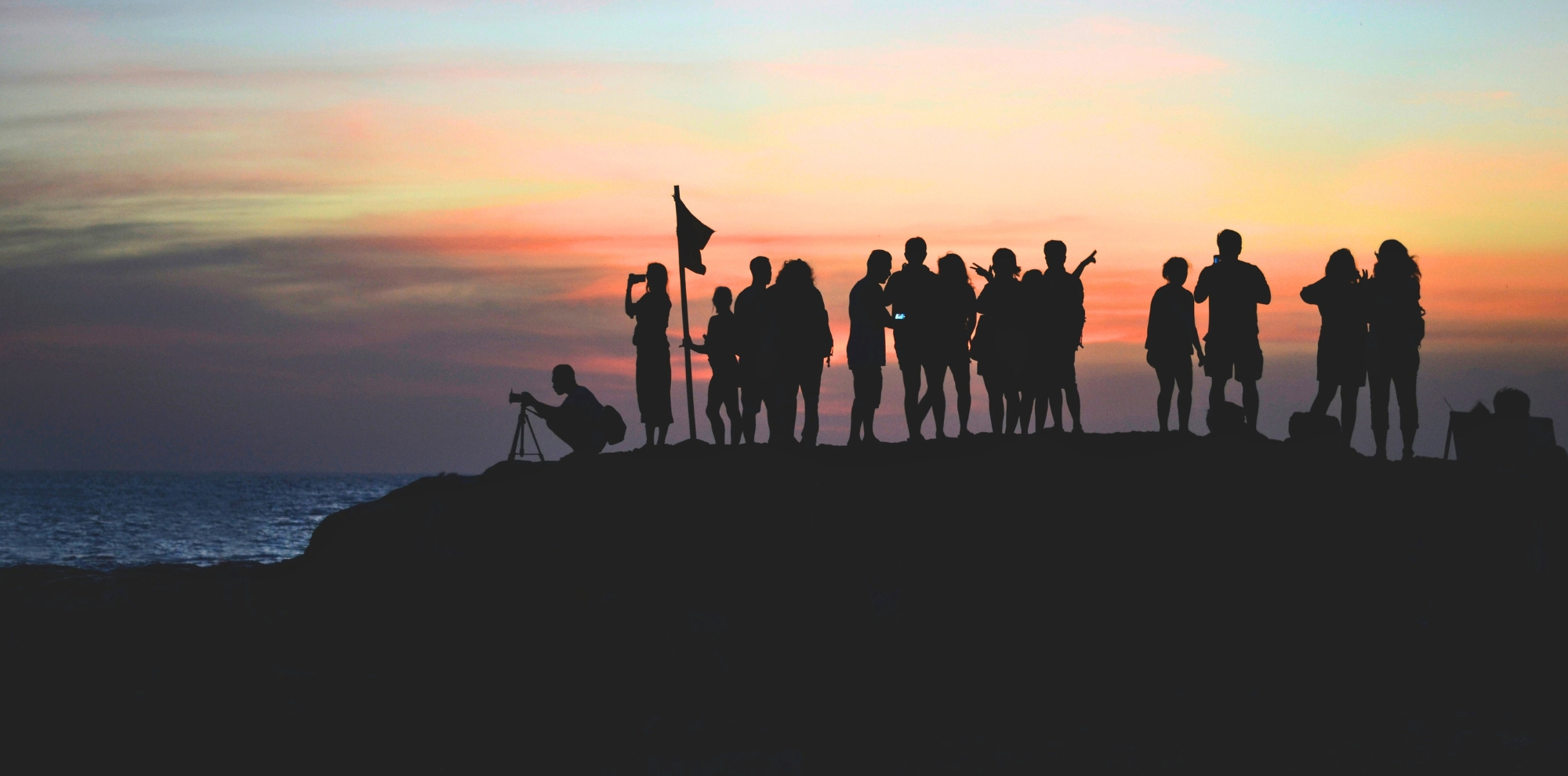 Silhouette of a group of people standing on a rocky cliff against a vibrant orange and pink sunset sky.