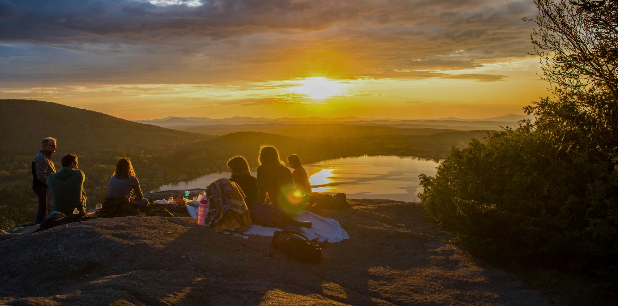 A group of friends sits on a rocky mountain summit, sharing a picnic while watching the sun set over a lake and distant hills.