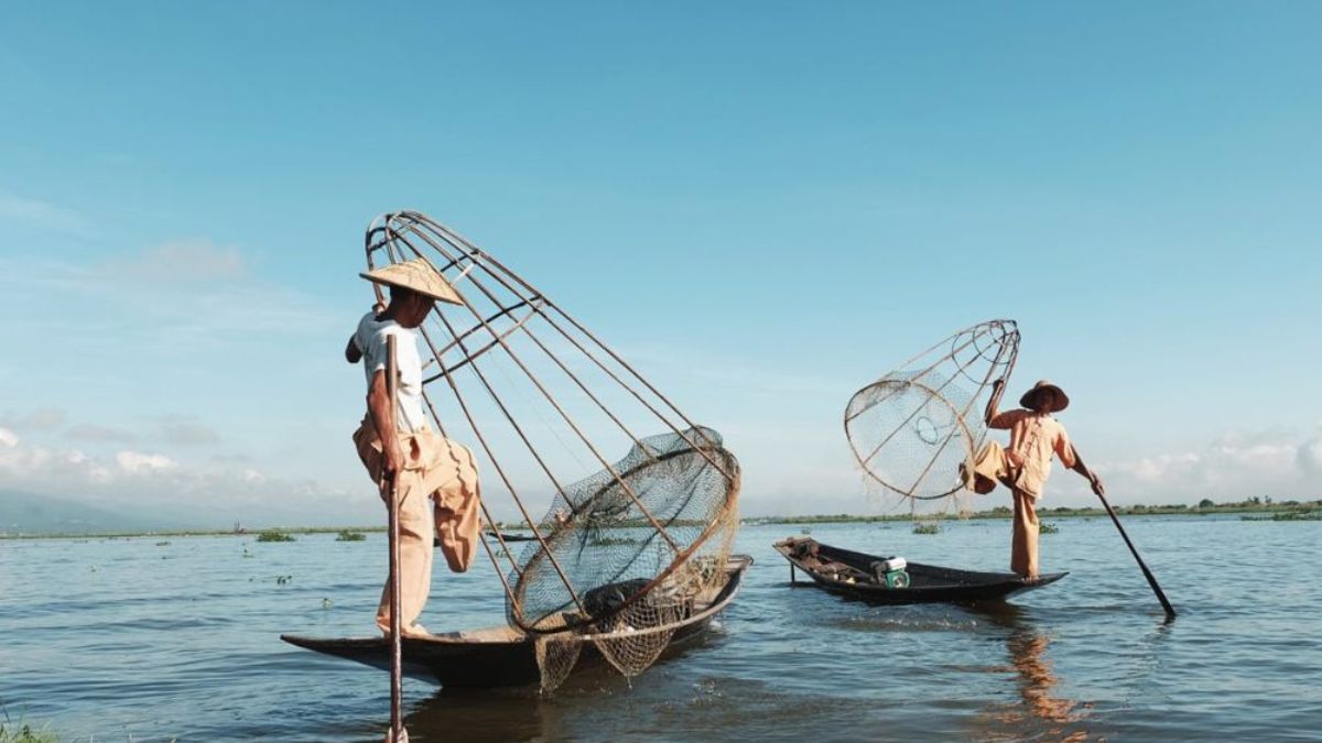 Two fishermen stand on narrow boats, balancing large conical nets while rowing with their legs on a calm lake.