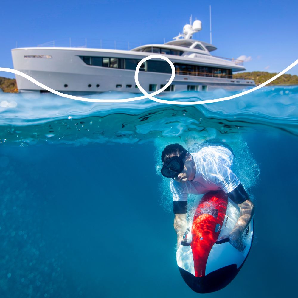 A man wearing goggles rides a red surfboard beneath the water’s surface, with a luxury yacht visible above him. The sky is clear and the water is a vibrant blue.