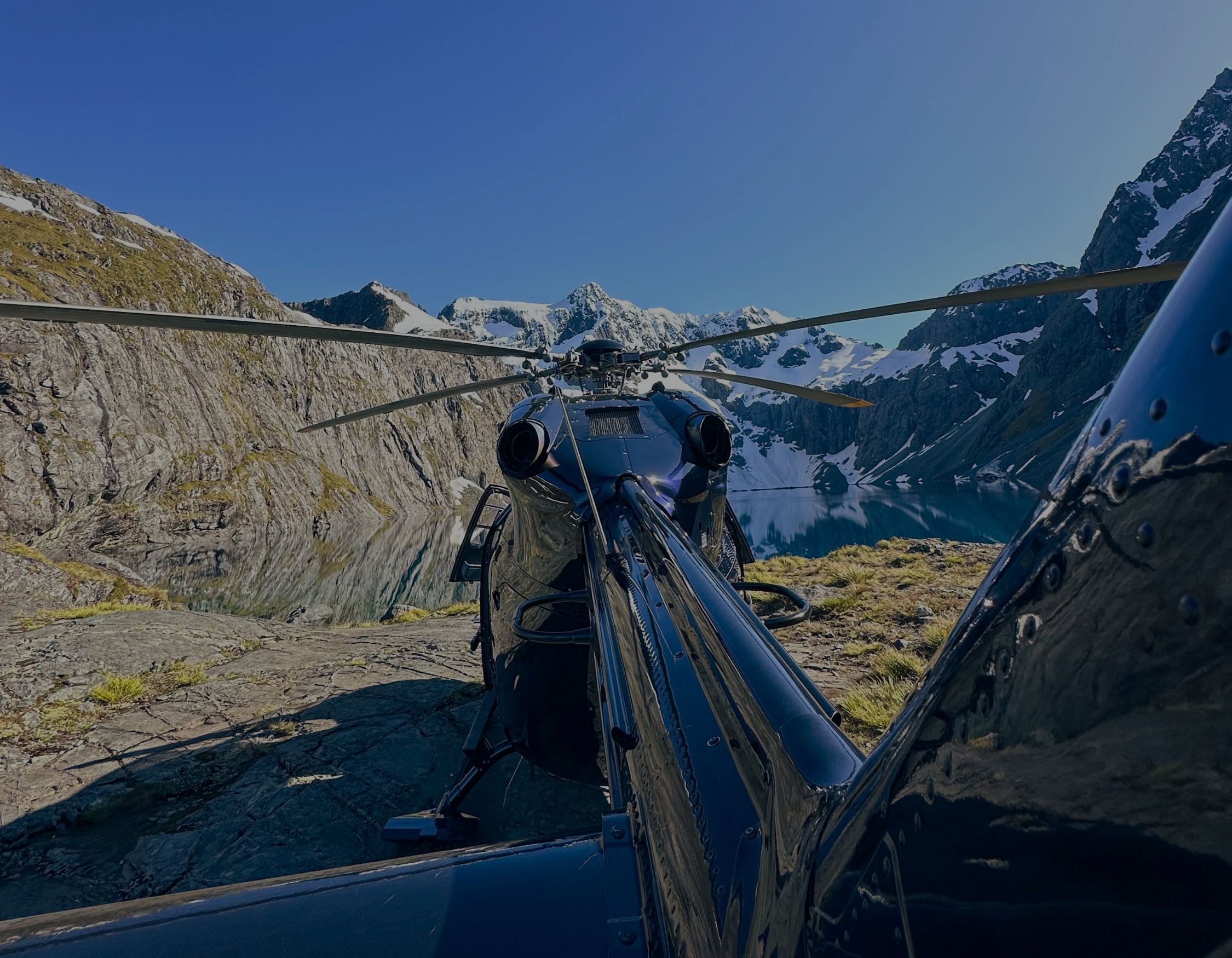 Rear view of a dark helicopter parked on a rocky ridge overlooking a mountain lake and snow-capped peaks.