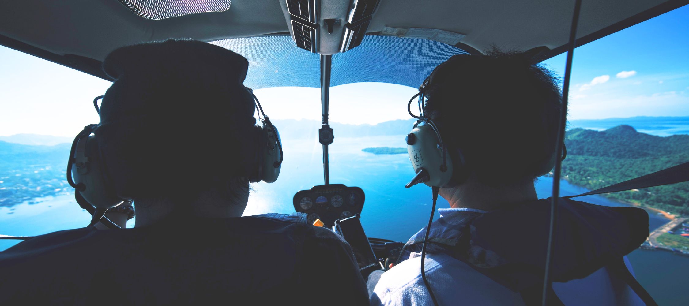 Two pilots wearing headphones are seen from behind, focused on flying a helicopter over a scenic landscape of water and islands. The sky is clear with few clouds, showcasing a bright and sunny day.