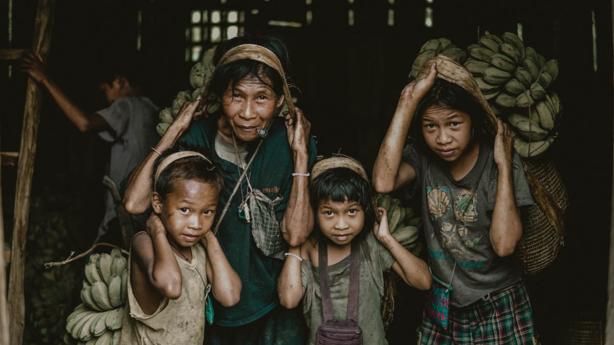 A group of four people, including two children and two women, carrying large baskets of bananas on their heads. They wear traditional clothing and look towards the camera with friendly expressions, against a darker background.