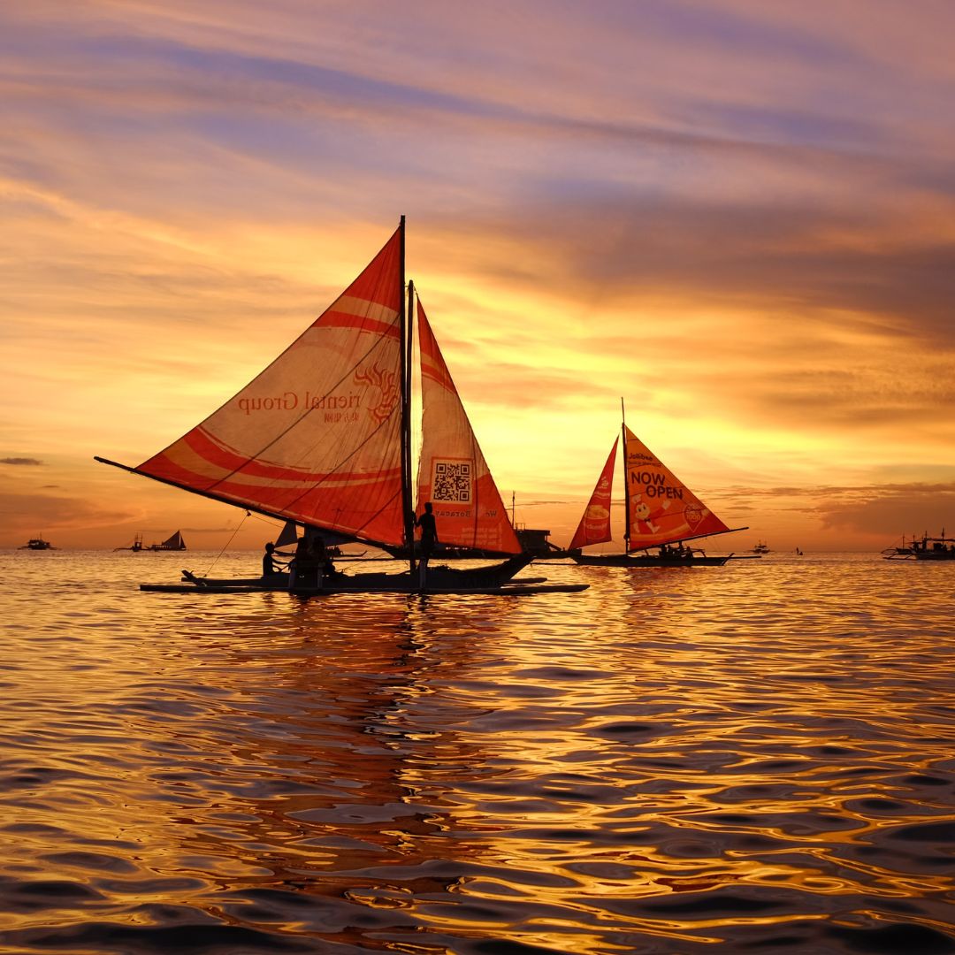 Sailboats with vibrant orange sails gliding over calm waters during a stunning sunset, reflecting warm hues of orange and purple in the sky.