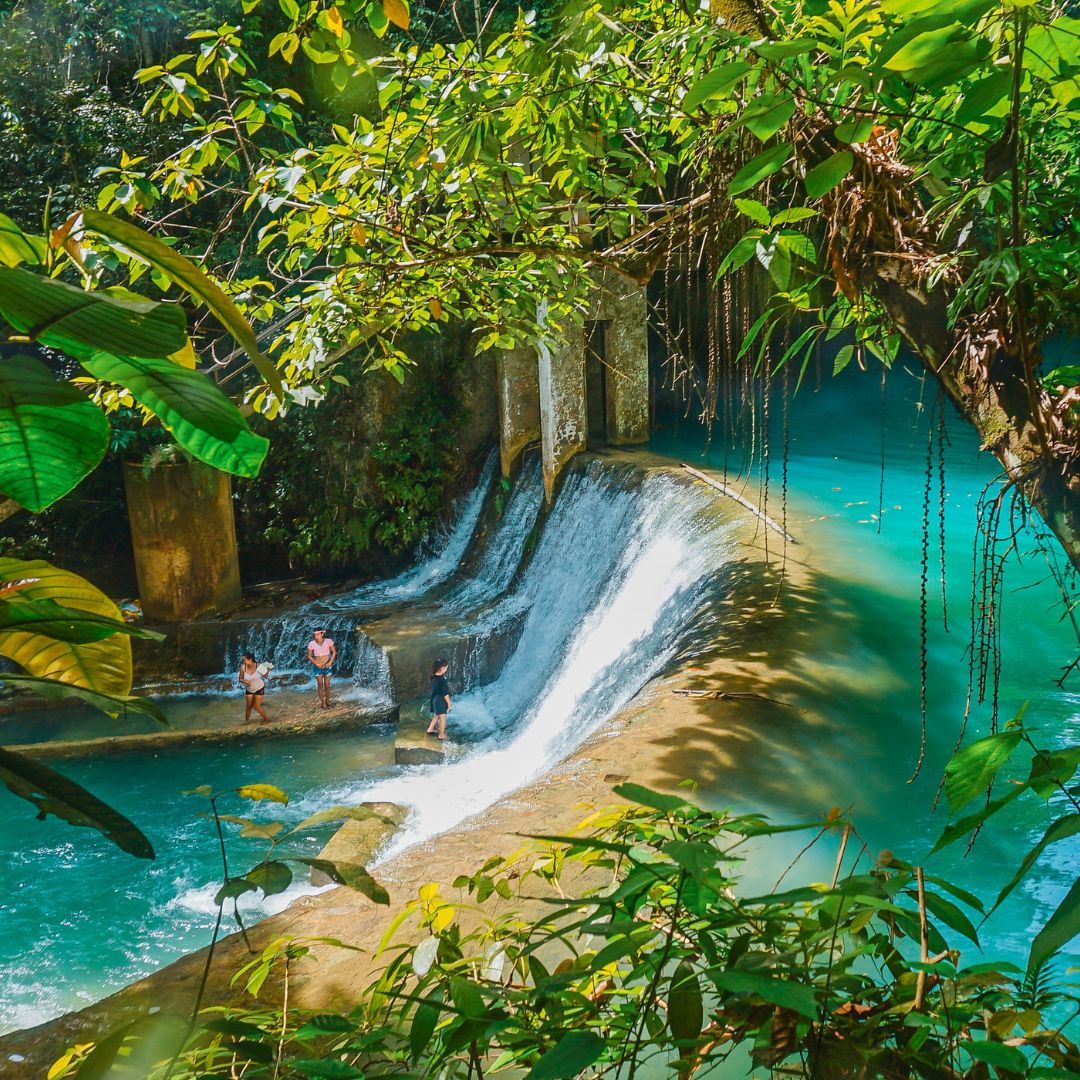 A bright blue waterfall cascades over a rocky ledge, surrounded by lush green foliage. Two people stand in the water, enjoying the serene natural setting.