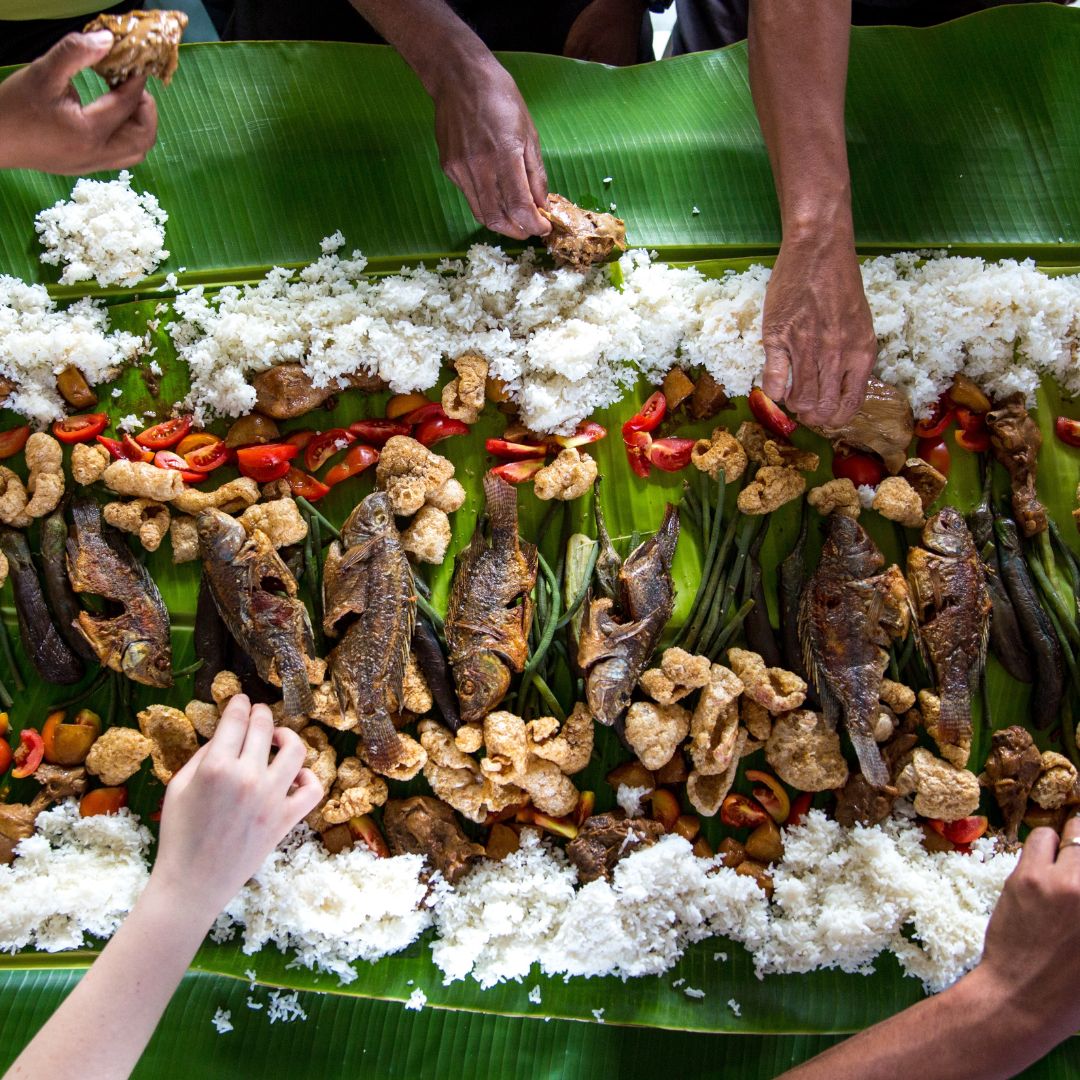 A communal feast on a large banana leaf, featuring grilled fish, fried plantains, and crispy snacks, surrounded by bowls of rice. Hands reaching in from different sides to share the meal.