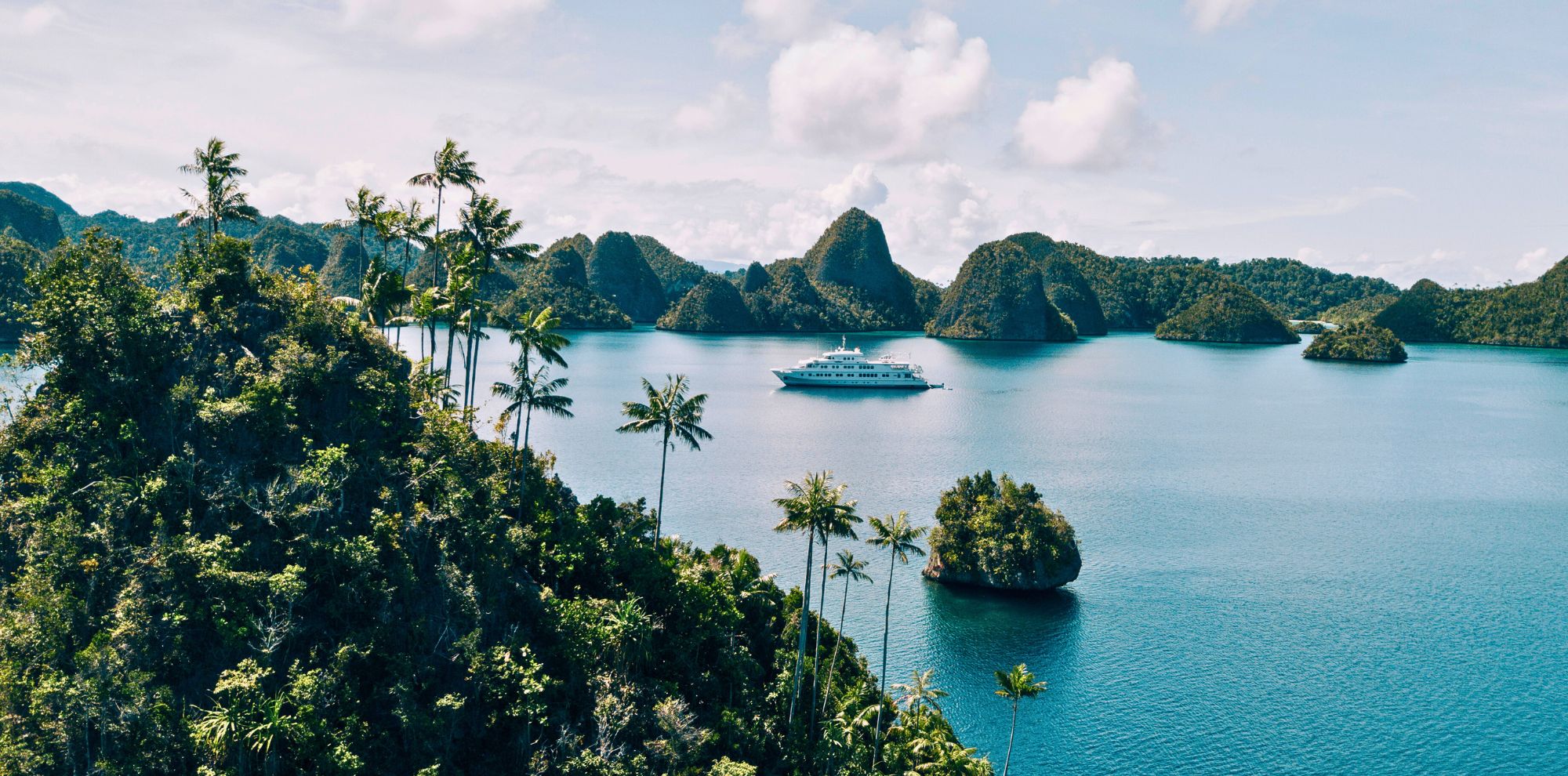 A luxurious yacht anchored in a tranquil blue bay surrounded by lush green islands and palm trees under a partly cloudy sky.
