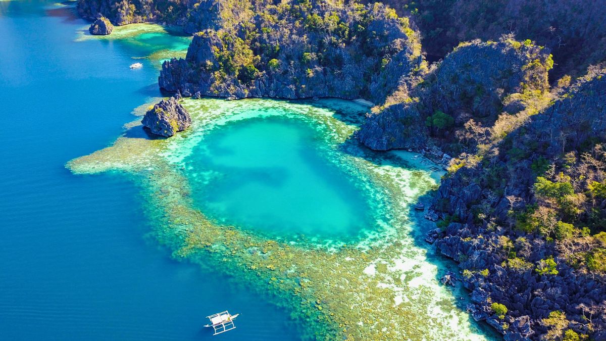 Aerial view of a turquoise lagoon surrounded by rocky cliffs and lush greenery, with a small boat floating on the clear water.