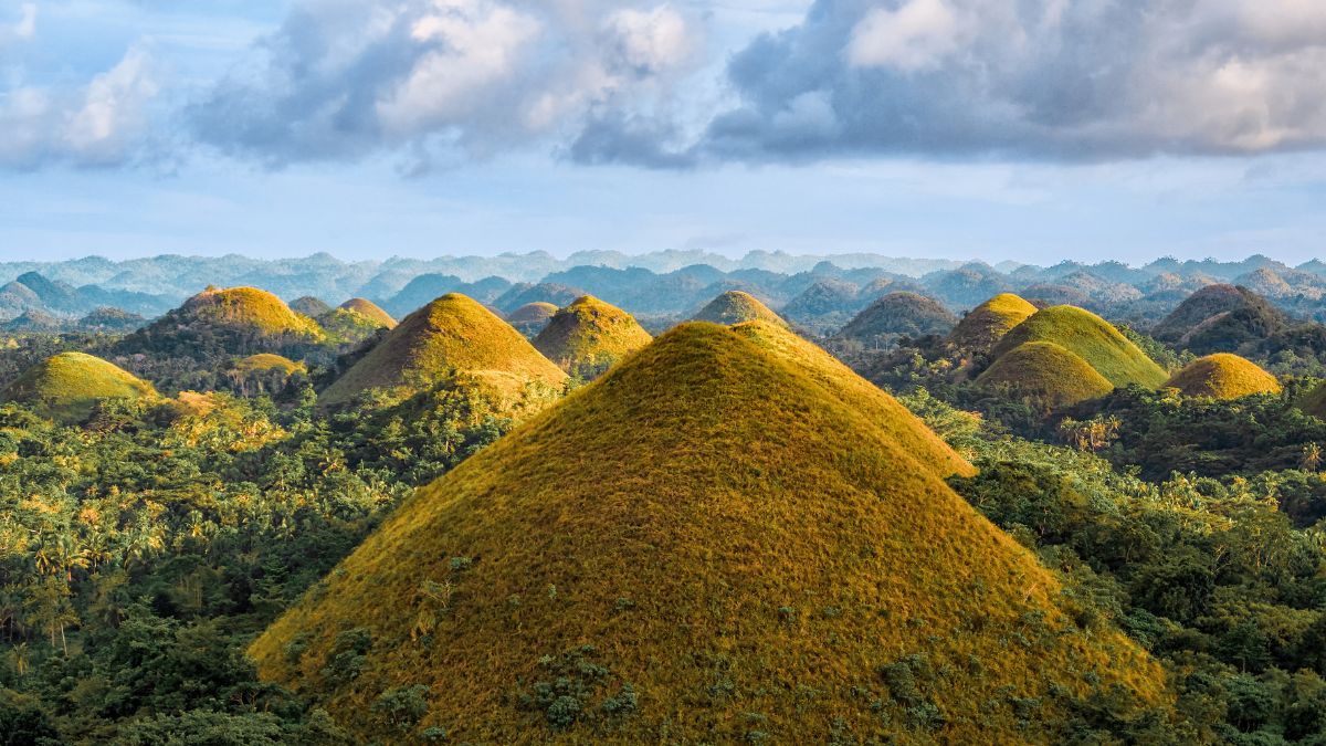 Lush, green hills shaped like conical mounds rise prominently against a blue sky with scattered clouds. The landscape showcases a vibrant array of greenery surrounding the formations.