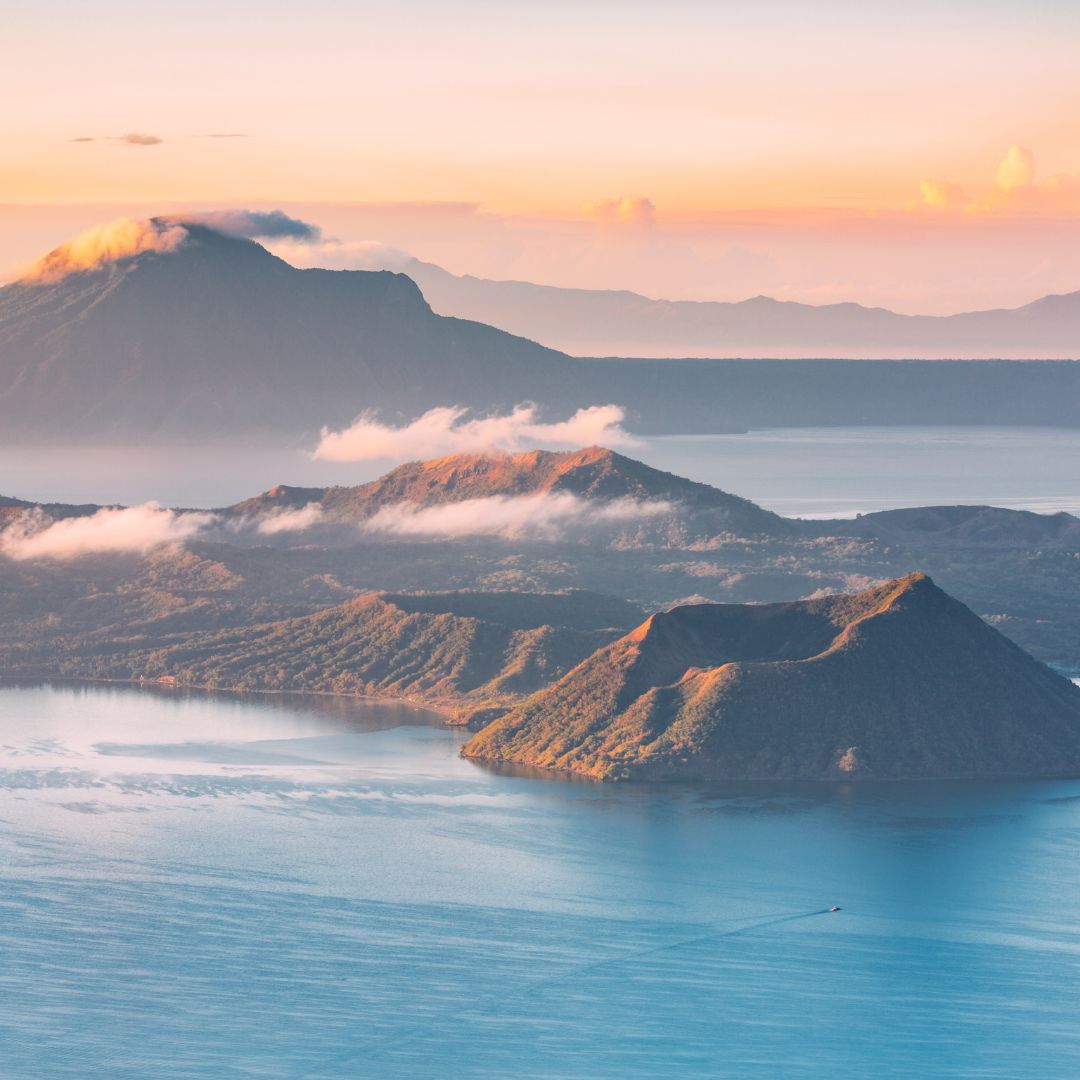 A serene landscape featuring a calm lake surrounded by lush green hills and distant mountains under a soft pastel sky. Wispy clouds drift above the peaks, illuminated by the warm hues of sunrise.