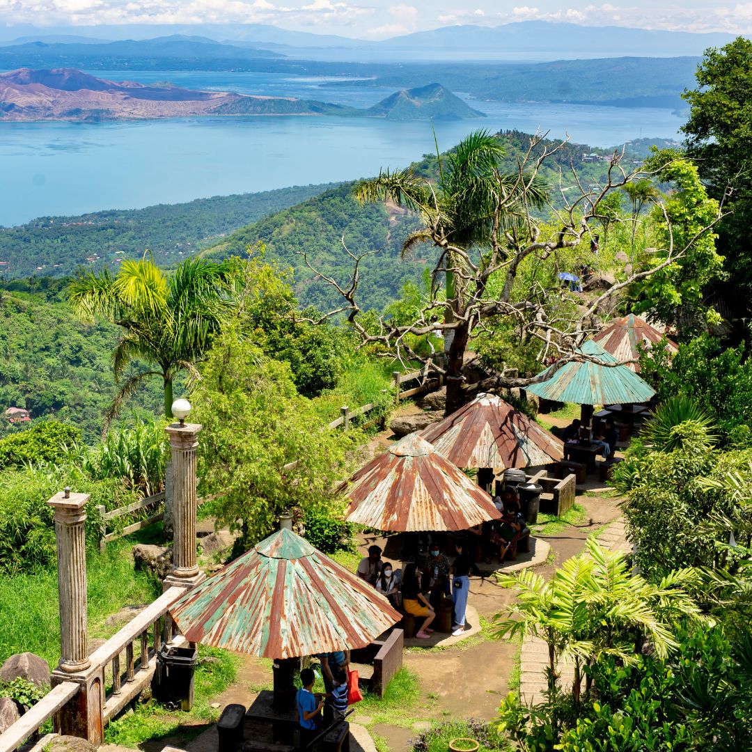 A scenic view overlooking Taal Lake, with green hills in the background and several rustic, corrugated iron-roofed huts shaded by palm trees. People are gathered in some of the huts, enjoying the outdoors.