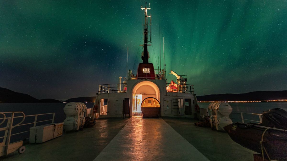 Green Aurora Borealis glows in a starry night sky above the deck of a ship with warm interior lighting.