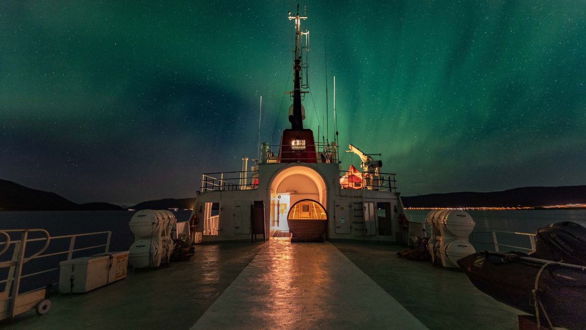 View from the deck of a ship at night, showing the glowing green aurora borealis streaking across a starry sky.