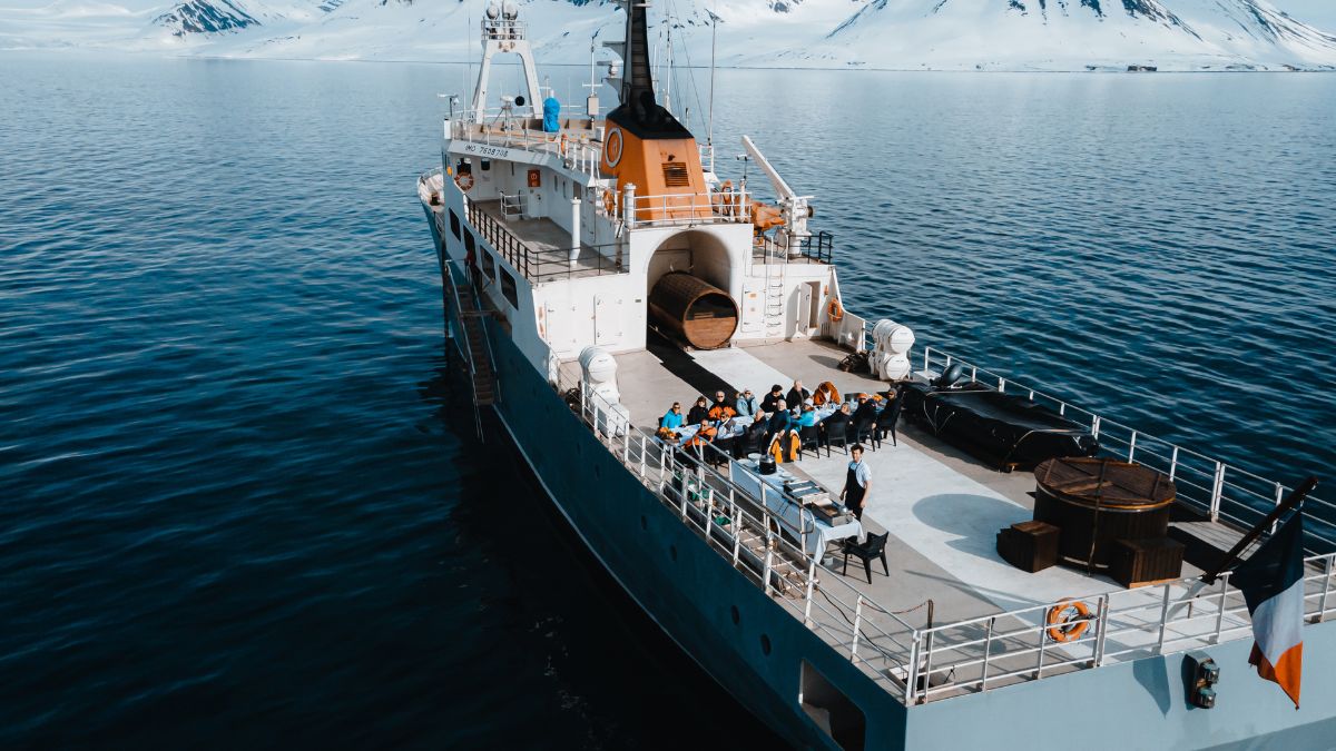 High-angle view of people dining at a long table on the deck of a ship in an arctic setting with snow-covered mountains.