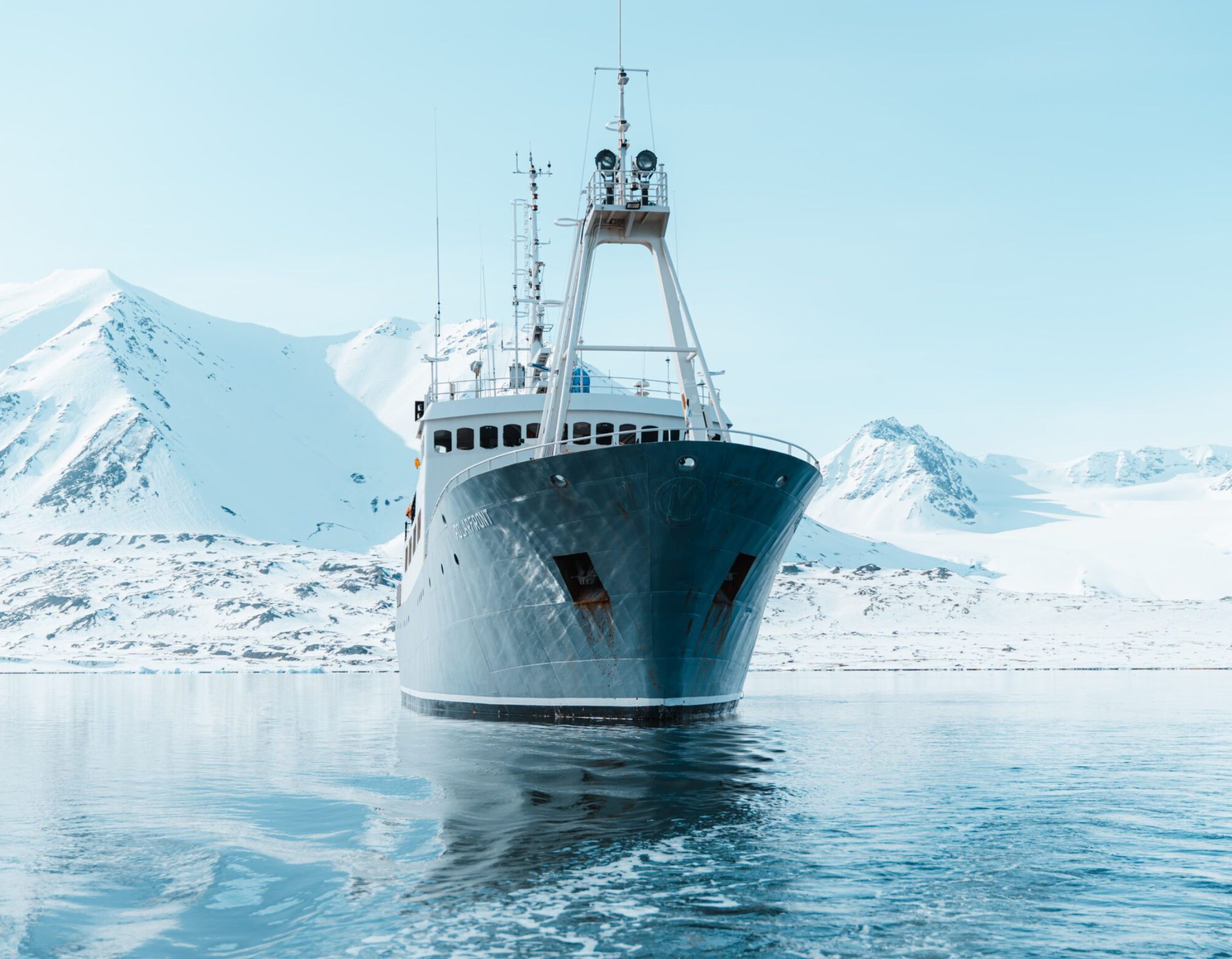 The bow of the ship POLARFRONT anchored in calm arctic waters with snow-covered mountains in the background.