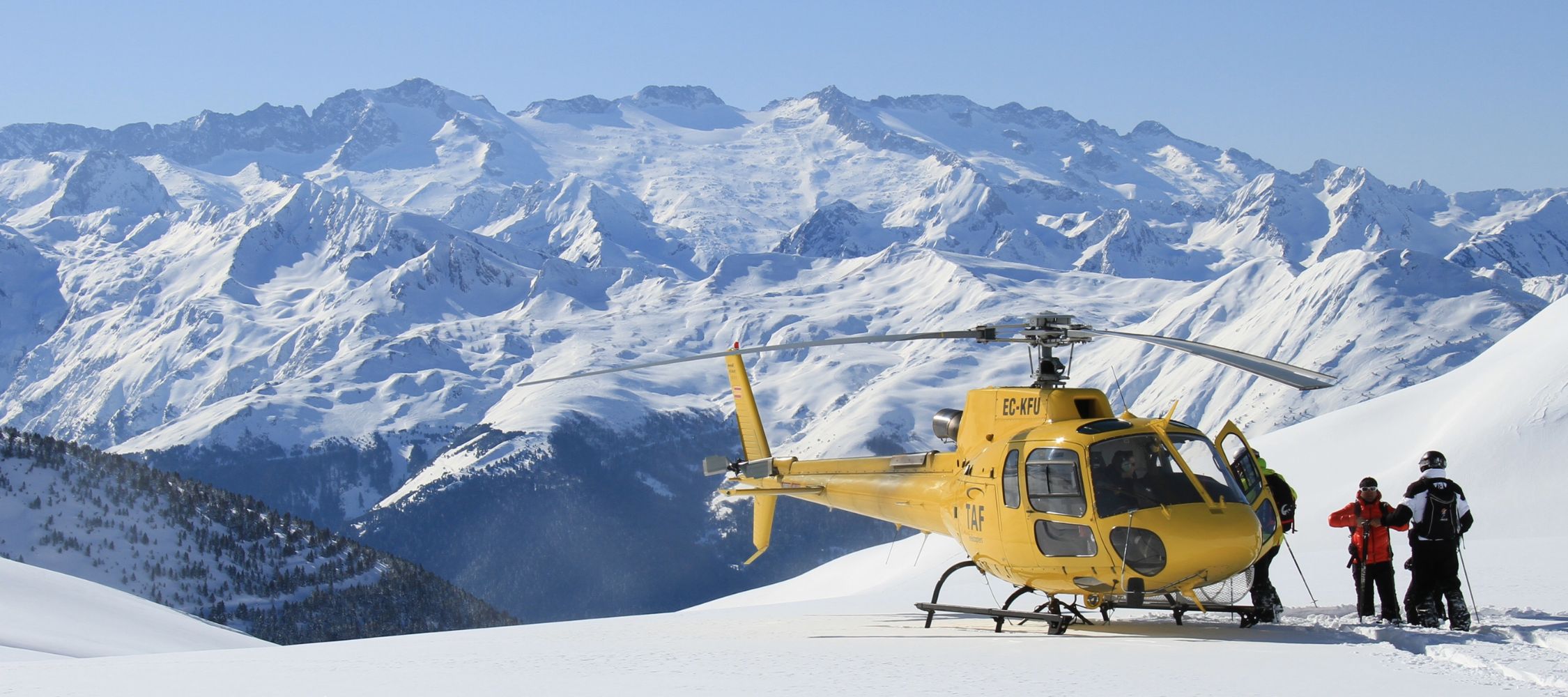 A yellow helicopter parked on a snowy mountain slope, surrounded by towering, snow-capped peaks under a clear blue sky. Two people in winter gear stand near the helicopter, enjoying the stunning alpine scenery.