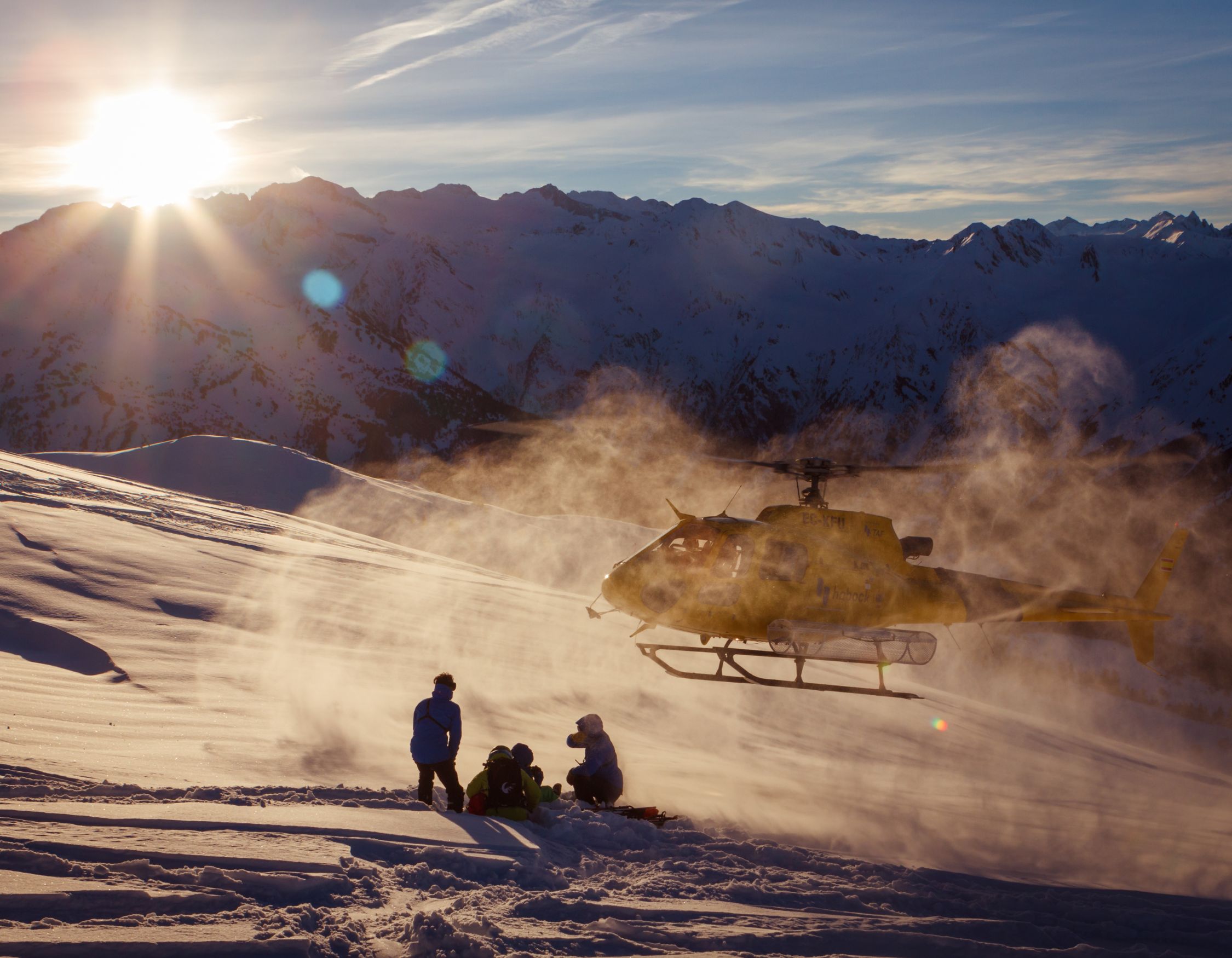 A helicopter lands on snow-covered mountain terrain as sunlight illuminates the scene. A group of three people is seated on the slope, with snow and mist swirling around them. In the background, majestic mountains rise under a clear blue sky.