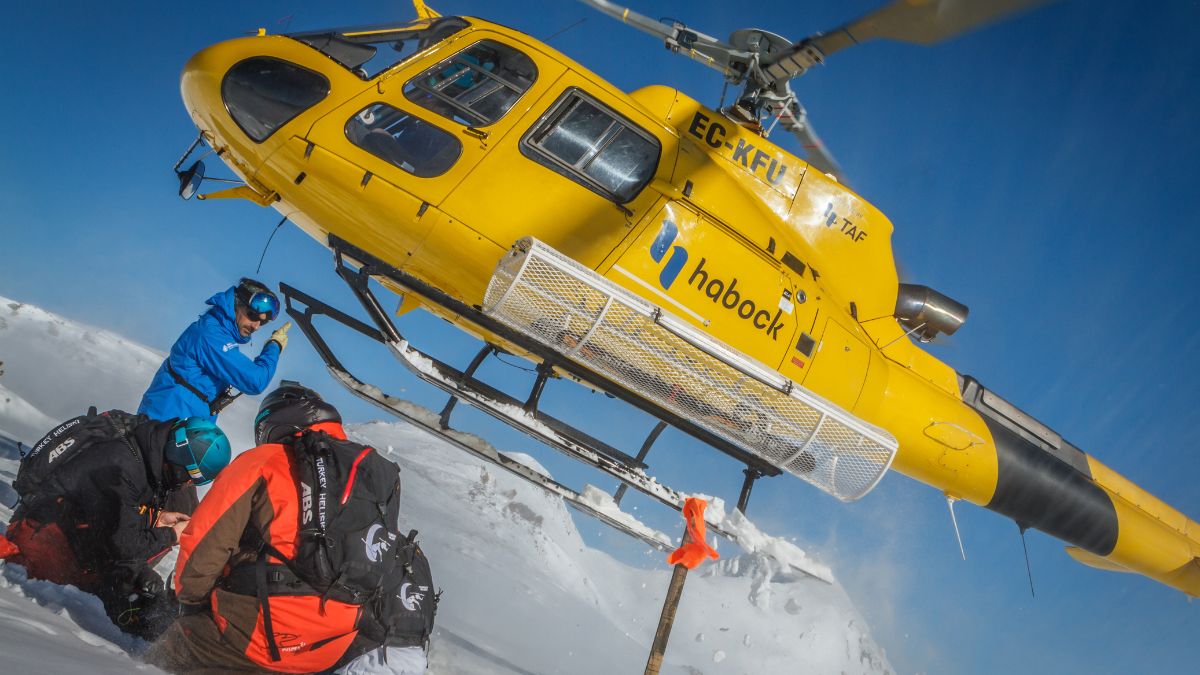 A bright yellow helicopter hovers above snowy terrain, with a rescue team dressed in winter gear attending to a person on the ground. The helicopter's landing gear is close to the snow, and the scene is set against a clear blue sky.