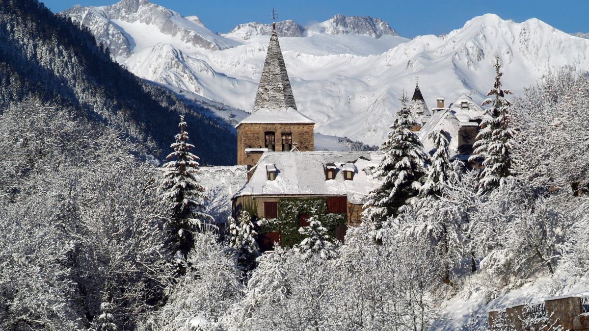 A quaint, snow-covered building with a tall spire is surrounded by frosted trees, set against a backdrop of majestic mountains.