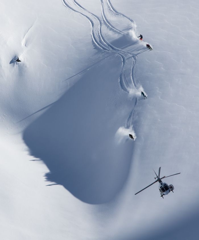 Helicopter hovering above skiers carving tracks in fresh snow on a mountainous slope.