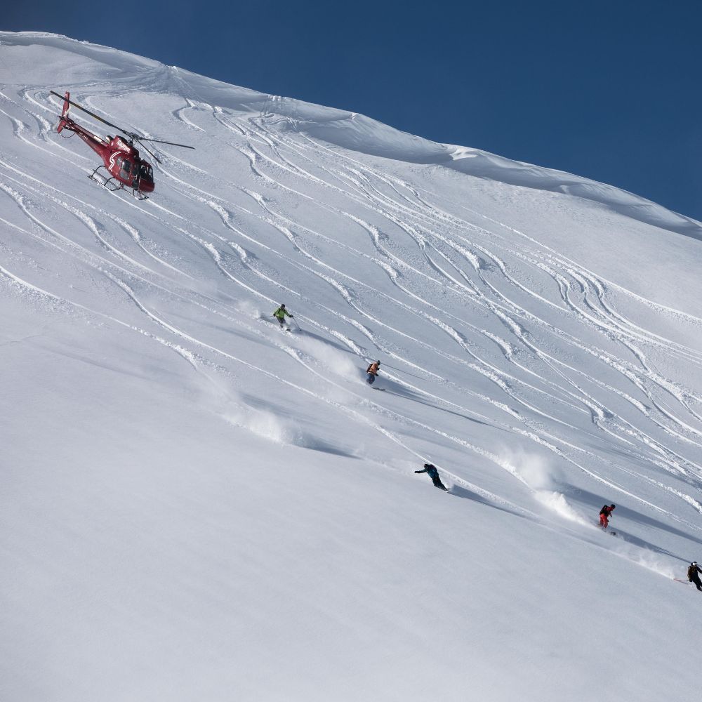 A red helicopter hovers above snowy slopes while several skiers carve tracks in fresh powder below. Clear blue sky in the background enhances the winter scene.