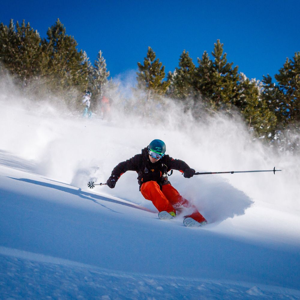 A skier in orange pants carves through fresh powder snow on a slope, with trees in the background and a clear blue sky overhead. Snow sprays up around them as they navigate the terrain.