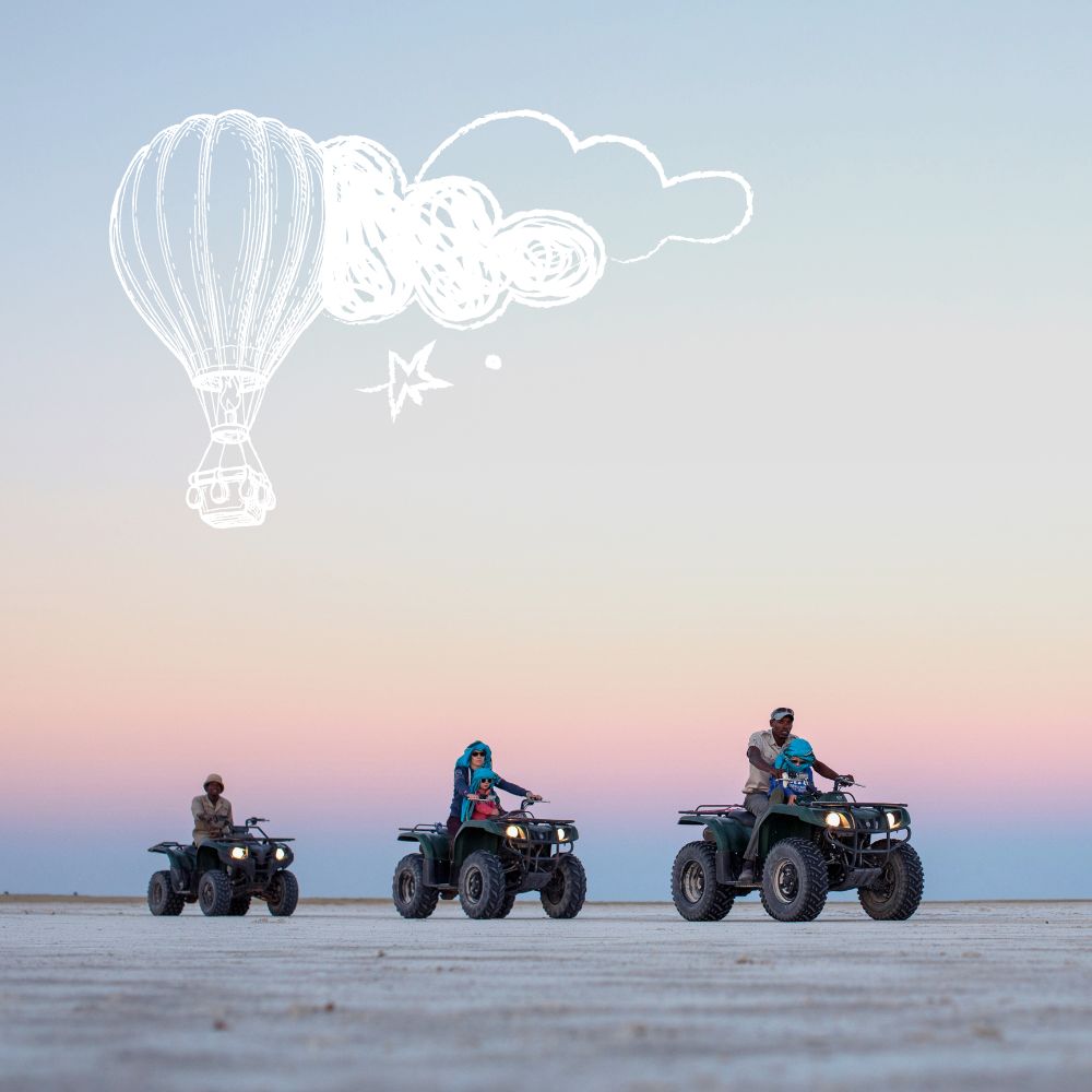 Three people ride all-terrain vehicles across a vast, flat landscape under a pastel sky. A whimsical hot air balloon and clouds are illustrated above them.