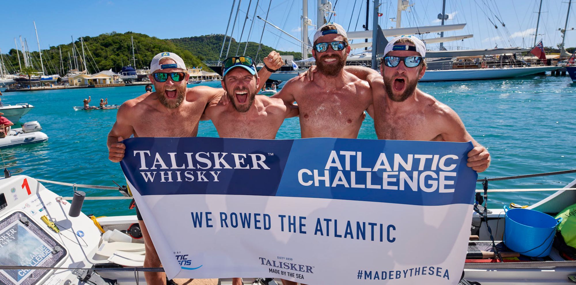 Four triumphant rowers pose shirtless on a boat, holding a banner that reads "Talisker Whisky Atlantic Challenge We Rowed the Atlantic." They celebrate their achievement against a backdrop of a marina and palm trees.